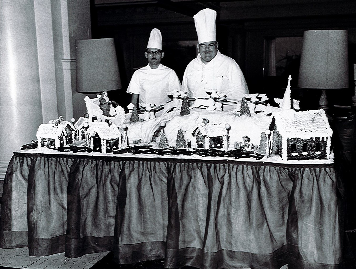 a couple of chefs standing next to a table with gingerbread houses