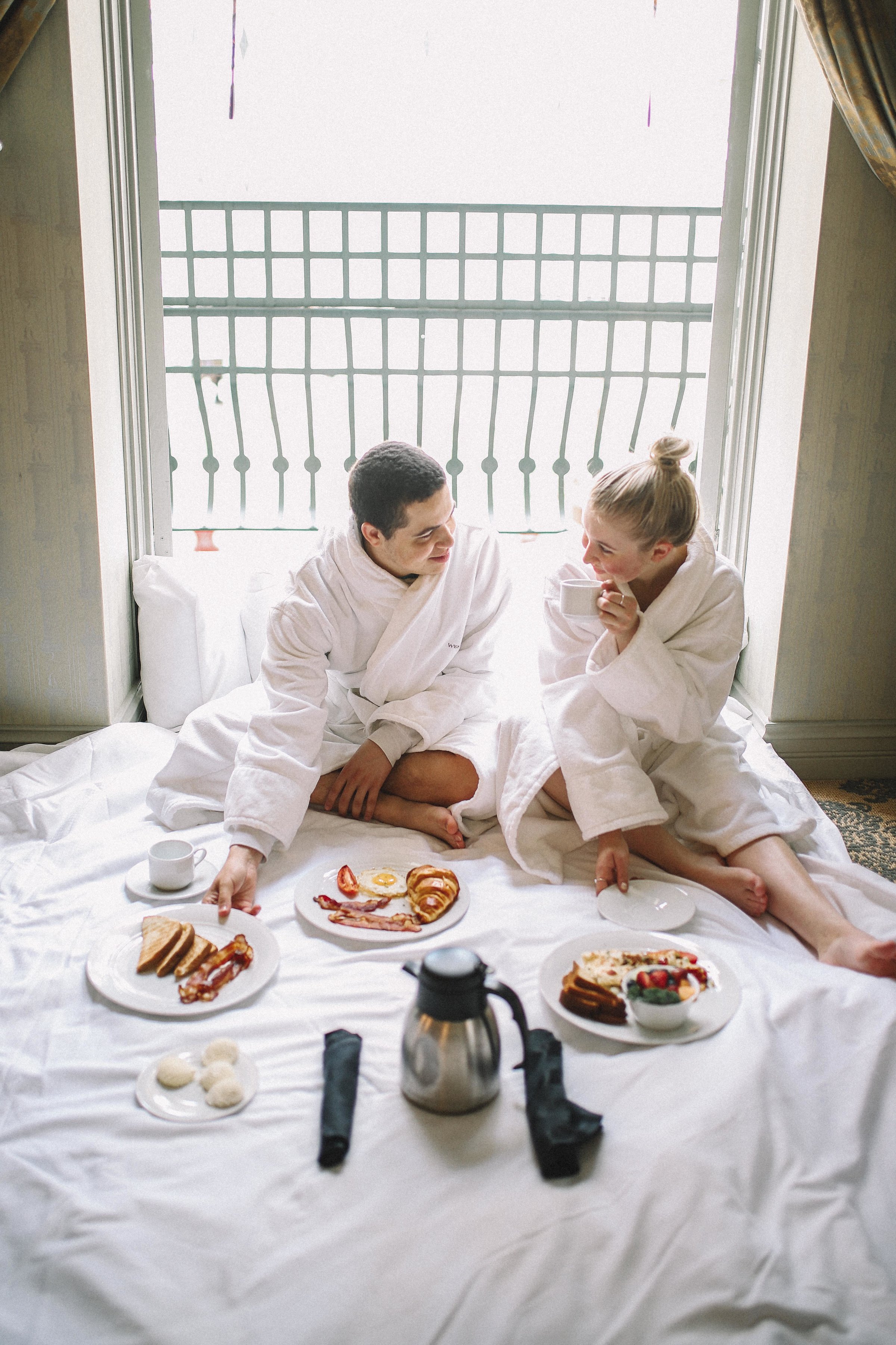 a man and woman in white robes sitting on a bed with plates of food